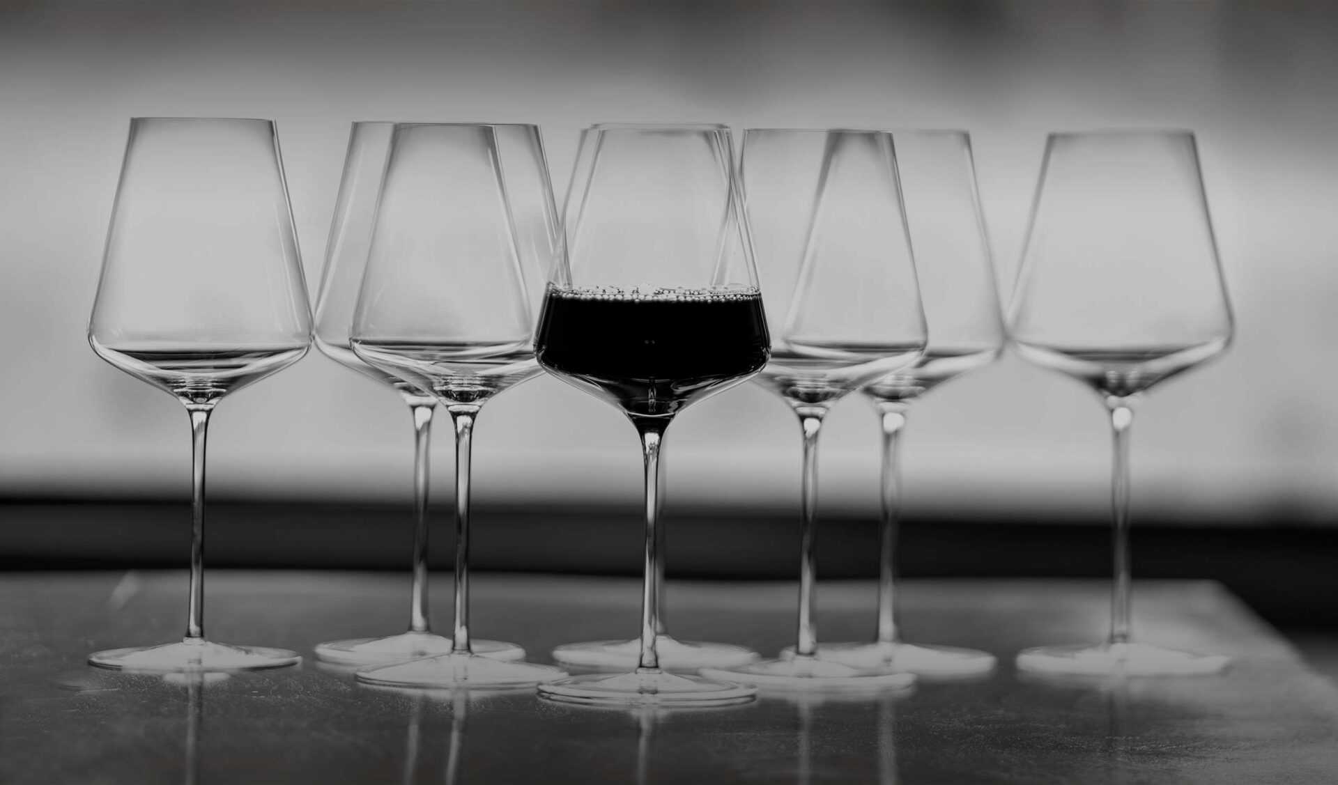 A set of seven wine glasses arranged in a row, with the center glass filled with red wine. The other glasses are empty. The image is in black and white, creating a contrast between the dark wine and the clear glasses.