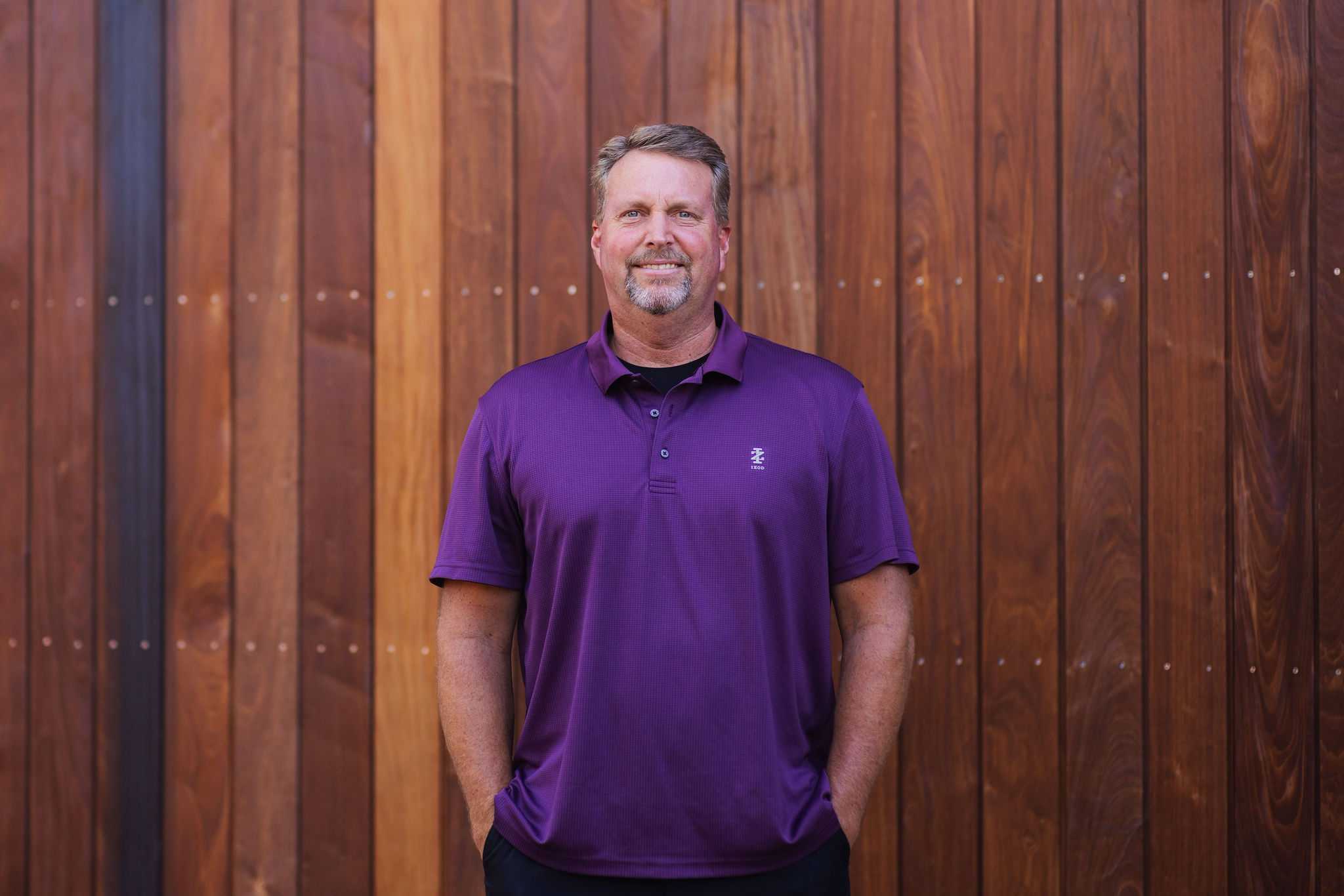 A man with short hair and a beard, wearing a purple polo shirt stands with his hands in his pockets. He is against a wooden panel wall with rich, warm tones.