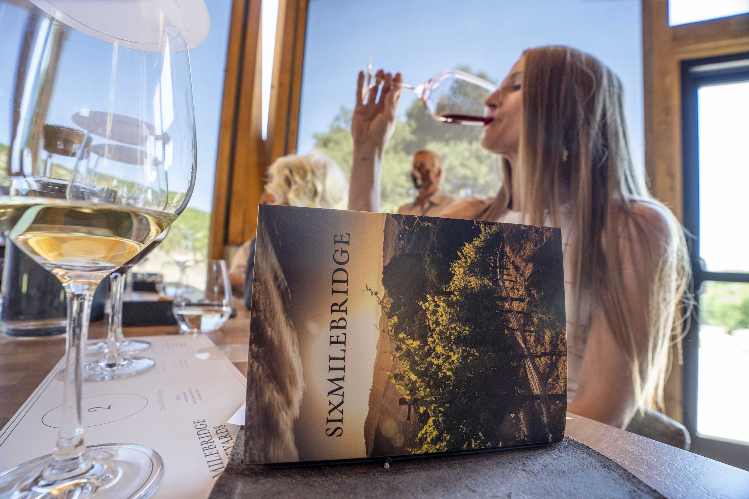 A woman with long hair sips wine at a table. In the foreground, a brochure with "Sixmilebridge" printed on it sits on the table next to a wine glass. The setting is indoors with large windows in the background.