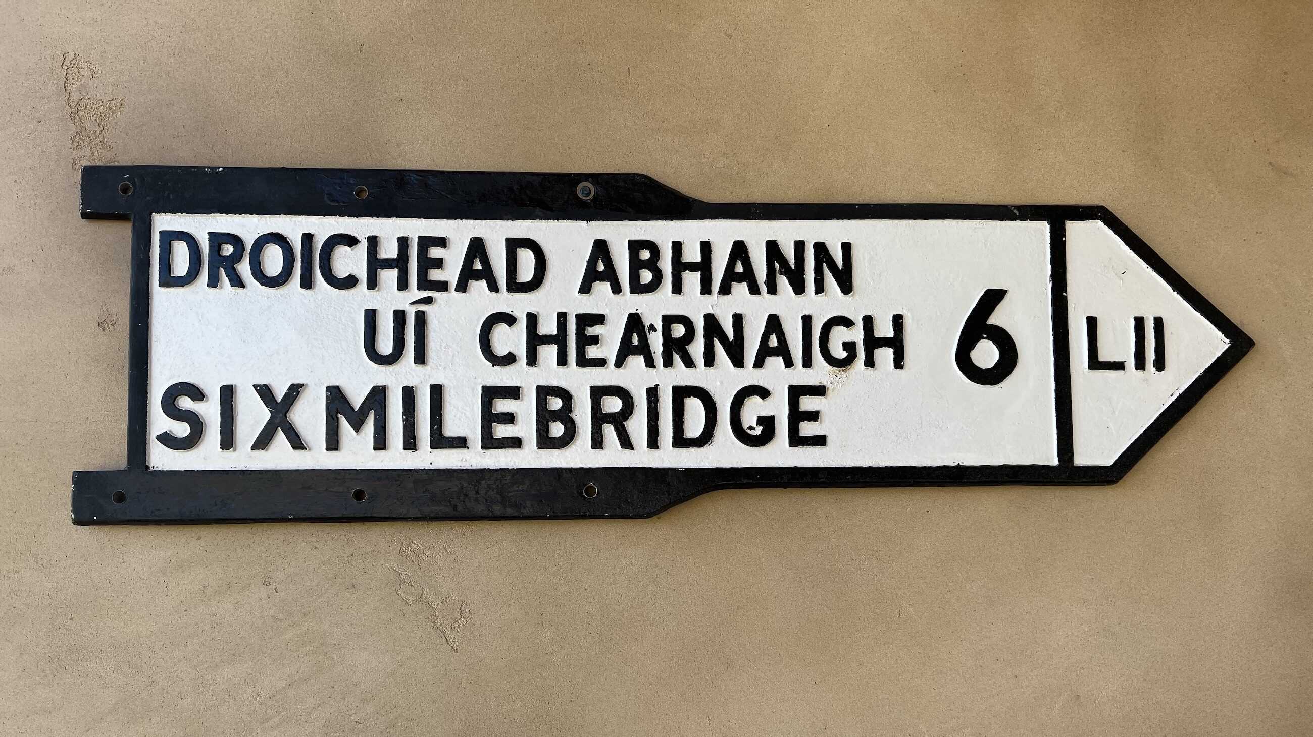 A vintage road sign with a black border on a beige background points to "Sixmilebridge" and its Irish name "Droichead Abhann Uí Chearnaigh," indicating a distance of six miles.