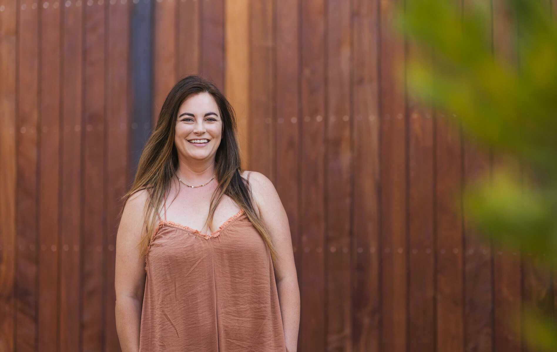 A woman with long brown hair stands smiling in front of a wooden wall. She is wearing a sleeveless brown top. The image has a warm and natural atmosphere.