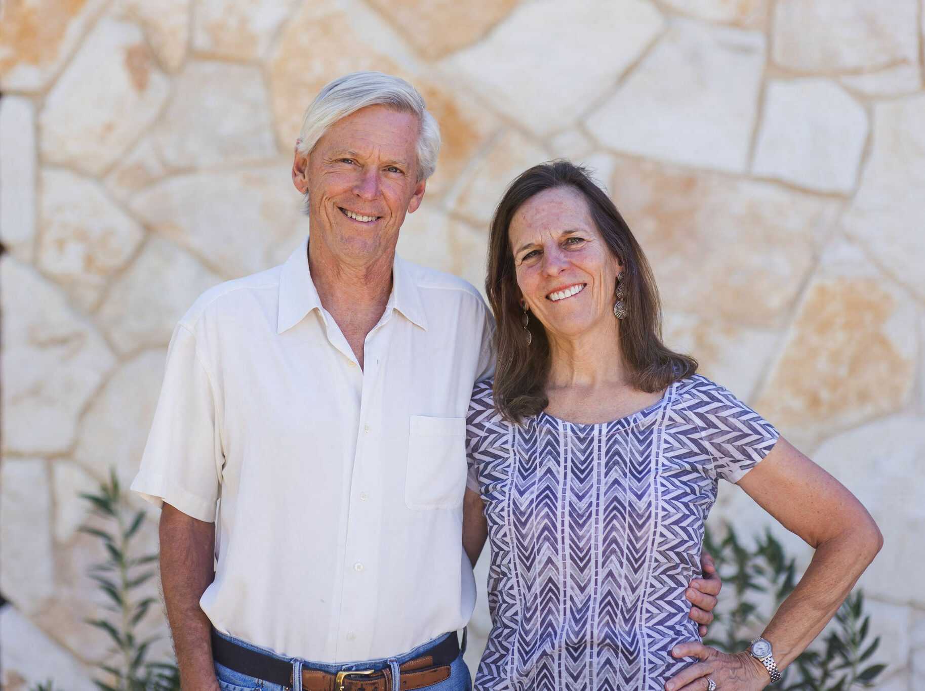 An older couple stands in front of a stone wall. The man, with gray hair, wears a white shirt and blue jeans. The woman, with long brown hair, wears a patterned dress. Both are smiling and appear relaxed.