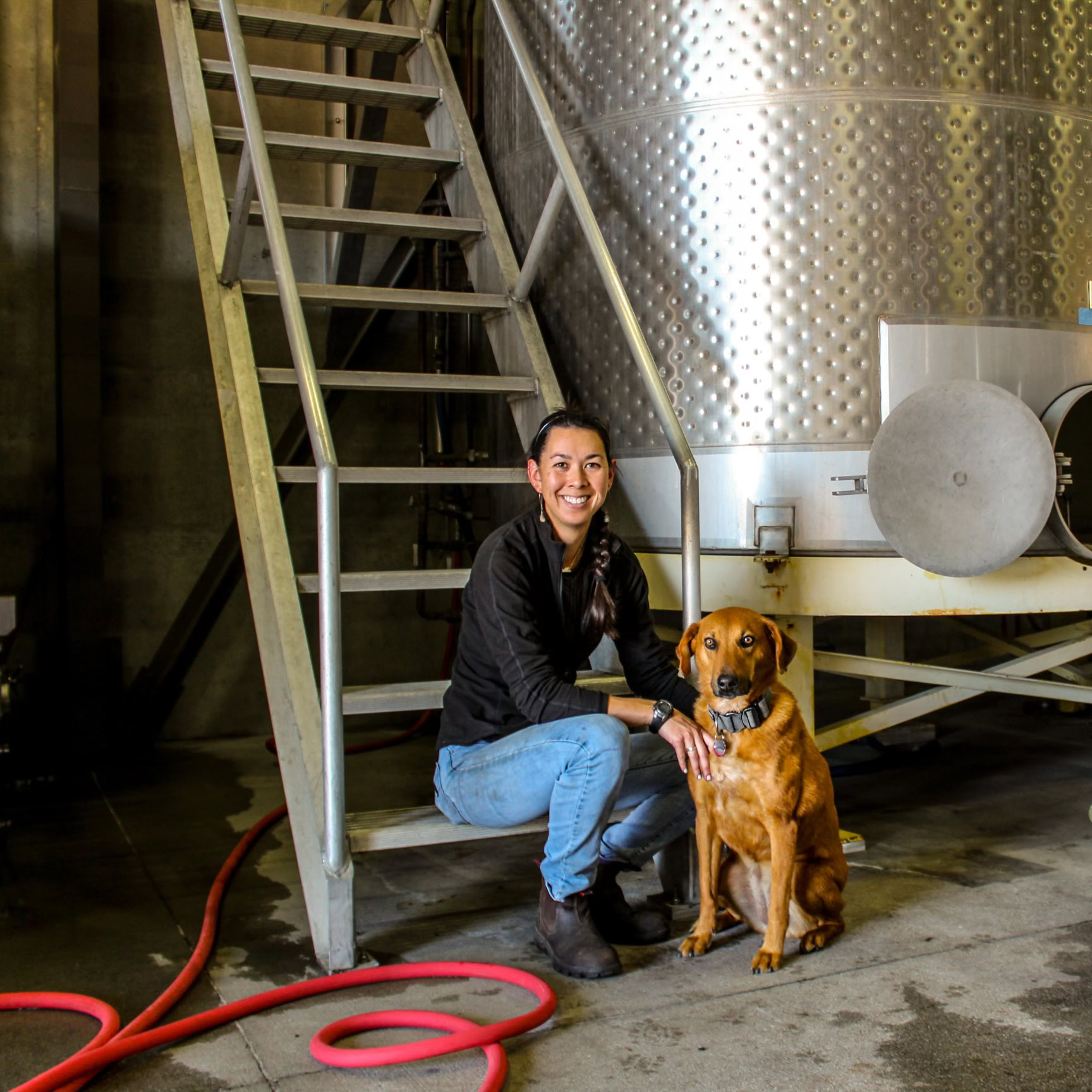 A person squatting beside a brown dog in an industrial setting with a large metal tank and a ladder in the background. Red hoses are visible on the ground.