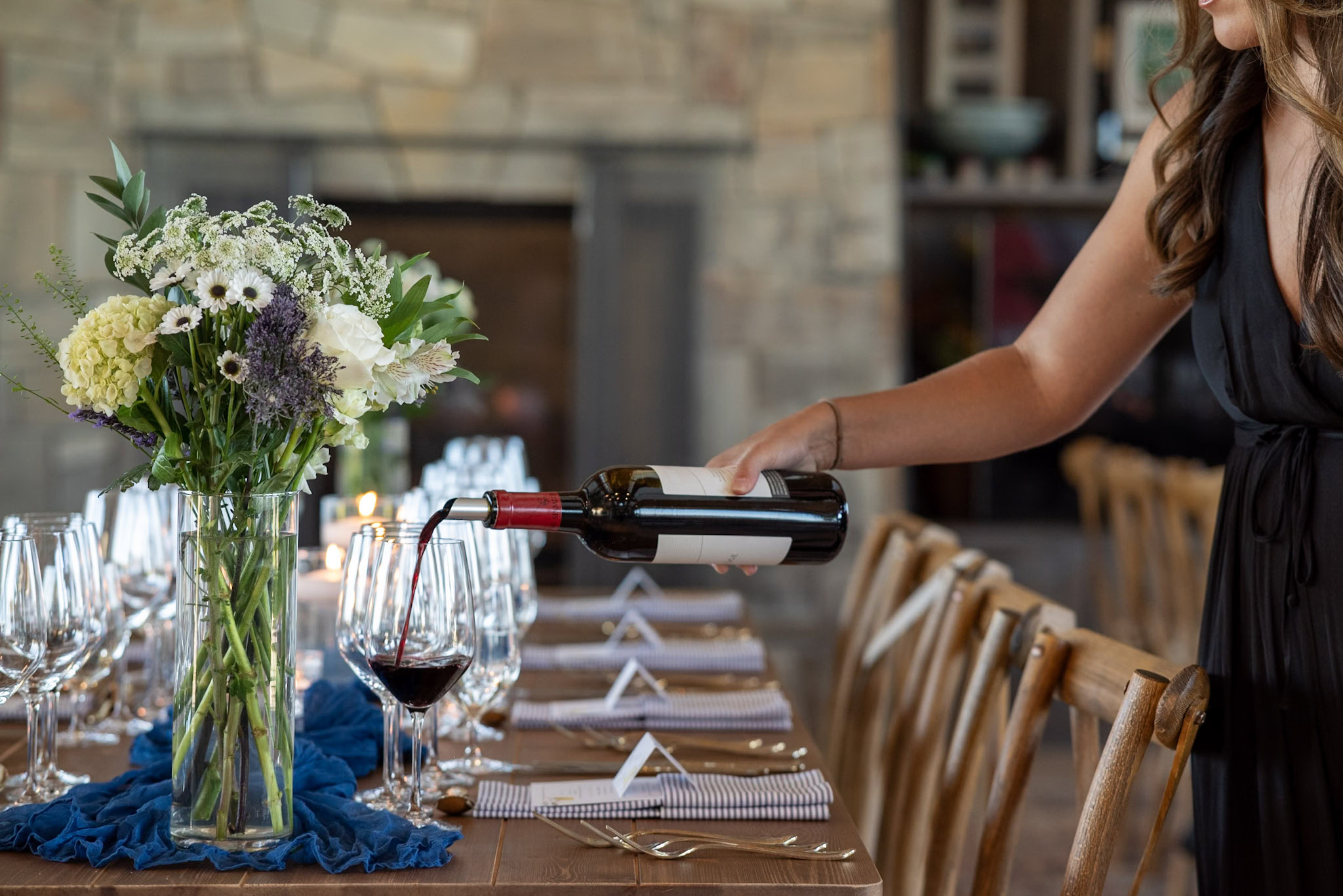 A person in a black dress pours red wine into a glass at a dining table set for a formal event. The table features elegant flower arrangements, candles, and neatly arranged glassware and cutlery. A stone fireplace is visible in the background.