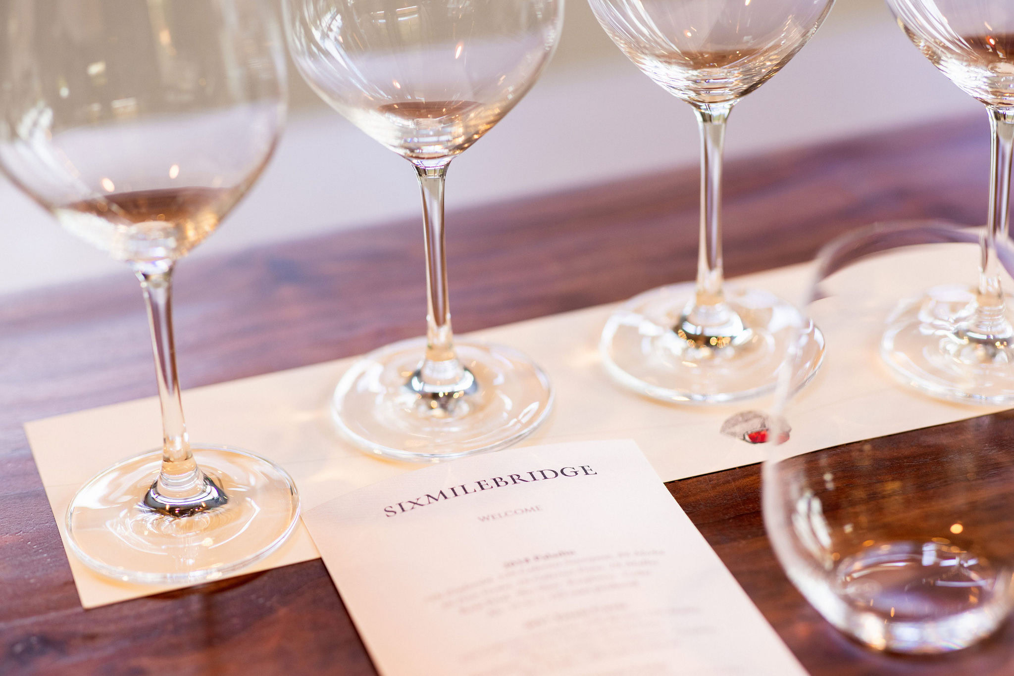 Four empty wine glasses on a wooden table, with a menu card from "Sixmilebridge" underneath, set for a tasting event.