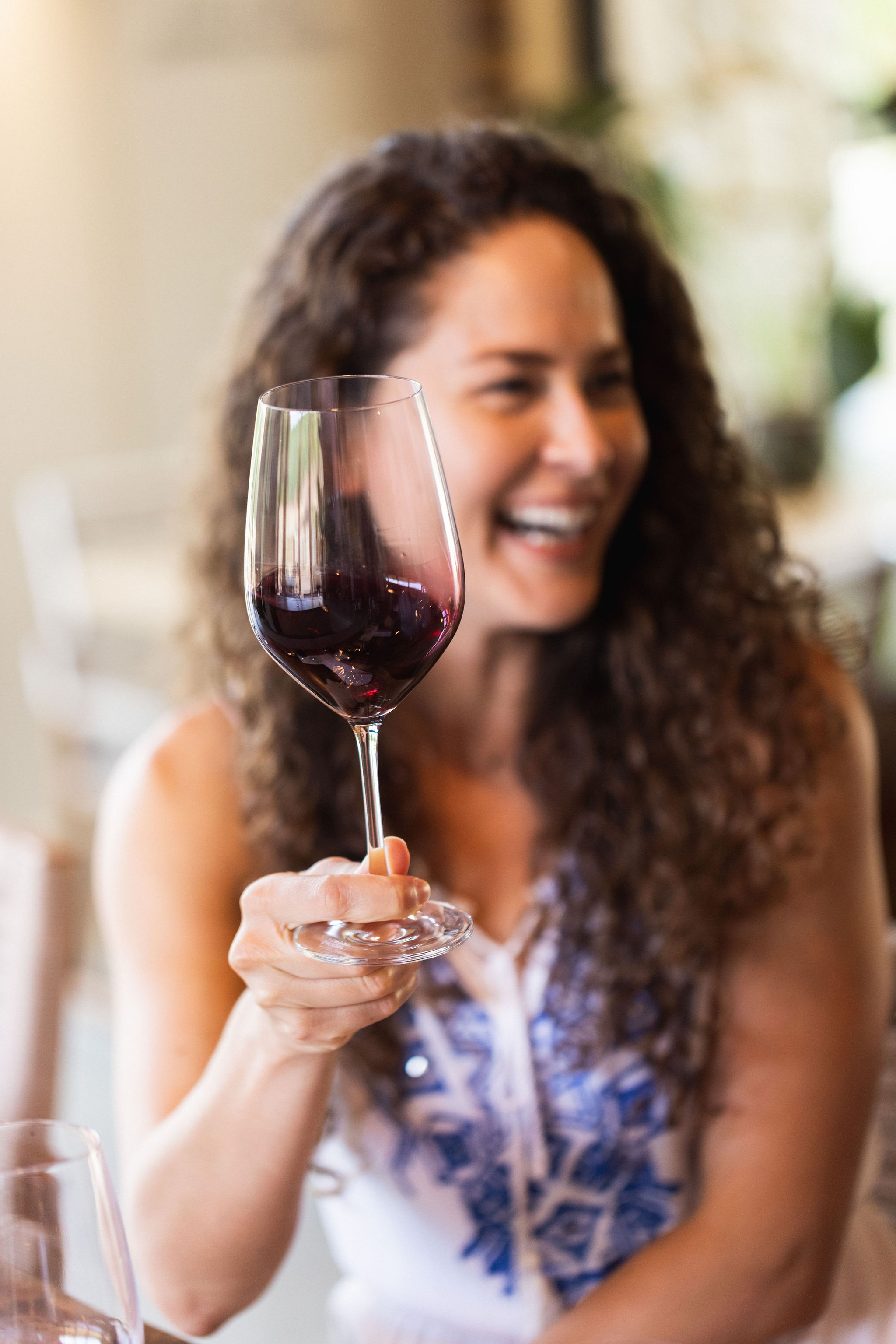 A woman with curly hair, smiling and holding a glass of red wine, is seated indoors. The background is softly blurred, giving a warm and inviting atmosphere.