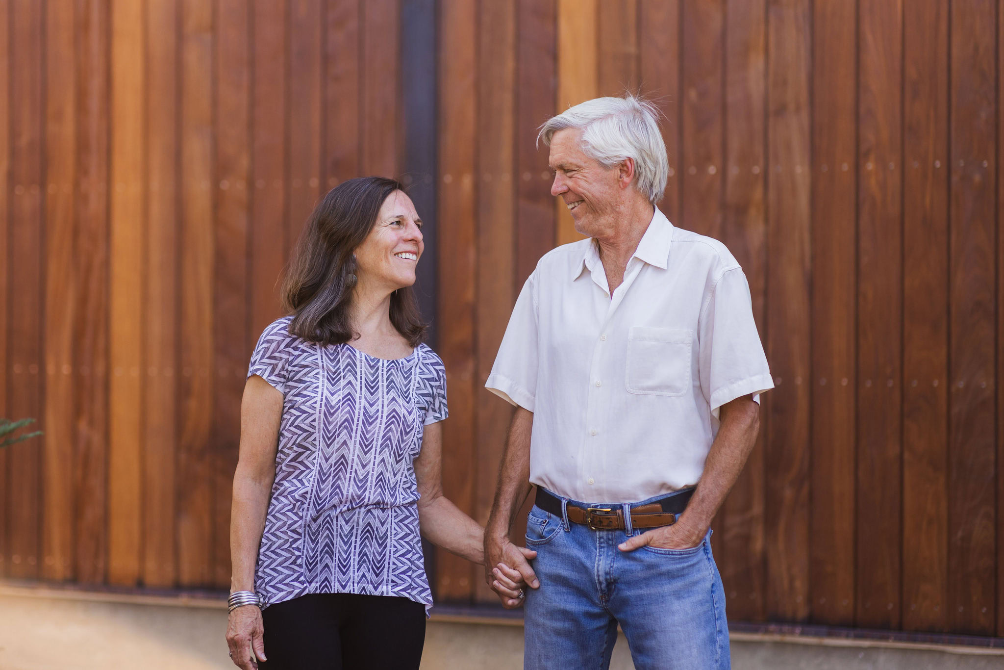 A smiling older couple holds hands, standing in front of a wooden wall. The woman has long brown hair and is wearing a patterned blouse, while the man has gray hair and is wearing a white short-sleeve shirt and jeans.