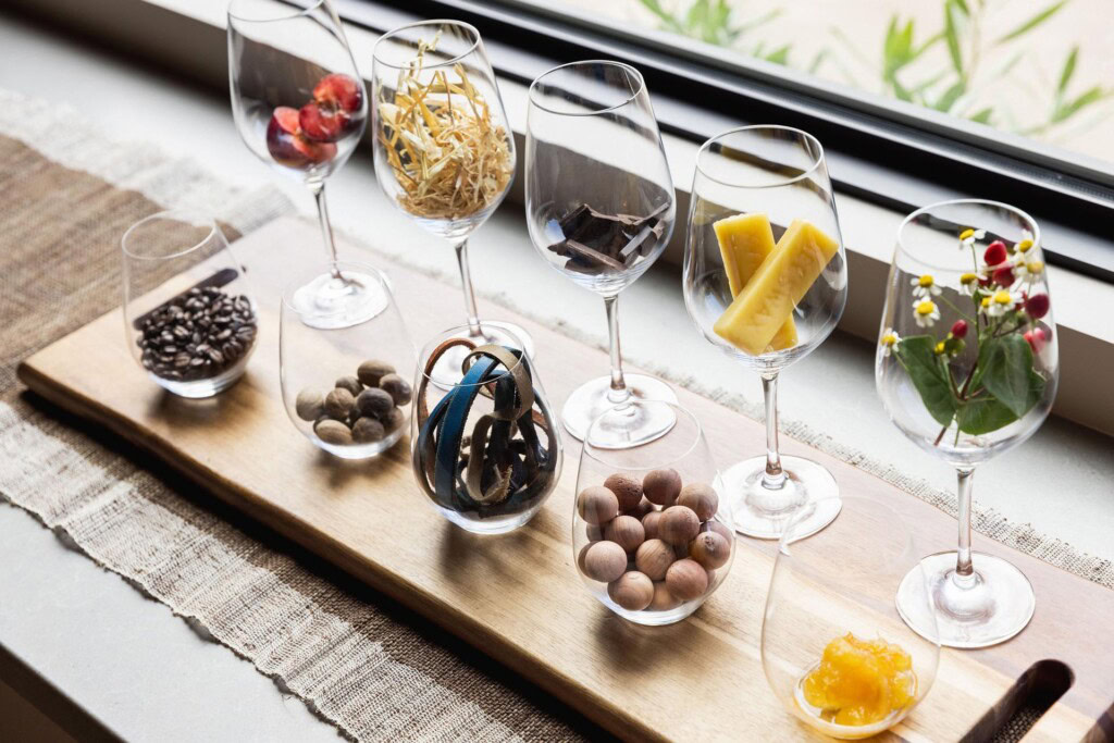 Wine glasses on a wooden board, each containing different items: coffee beans, nuts, dried fruit, spices, and flowers, arranged in a row. The setup is near a window with a neutral table runner underneath.