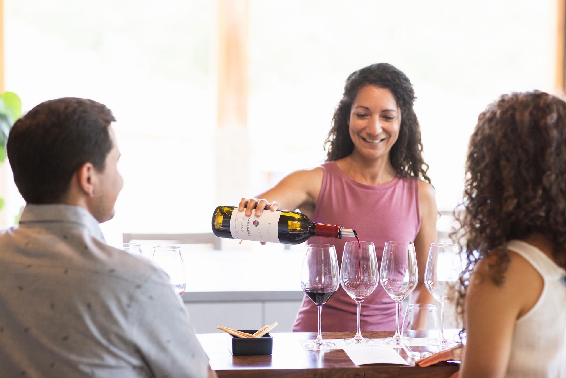 A woman in a pink sleeveless top pours red wine into a glass at a tasting session. Two people sit at the counter, facing her, with several empty and filled wine glasses in front of them. The setting appears bright and welcoming.