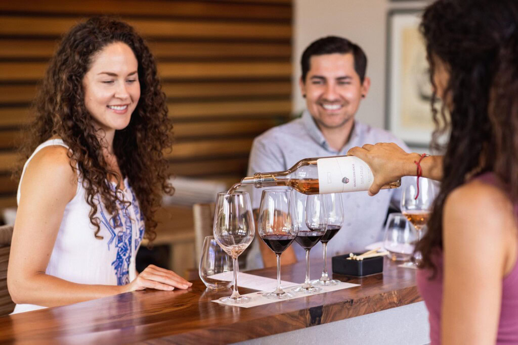 A person pours wine into a glass at a wooden counter while two people watch and smile. The setting appears to be a tasting room, with wine glasses and bottles arranged neatly on the counter.