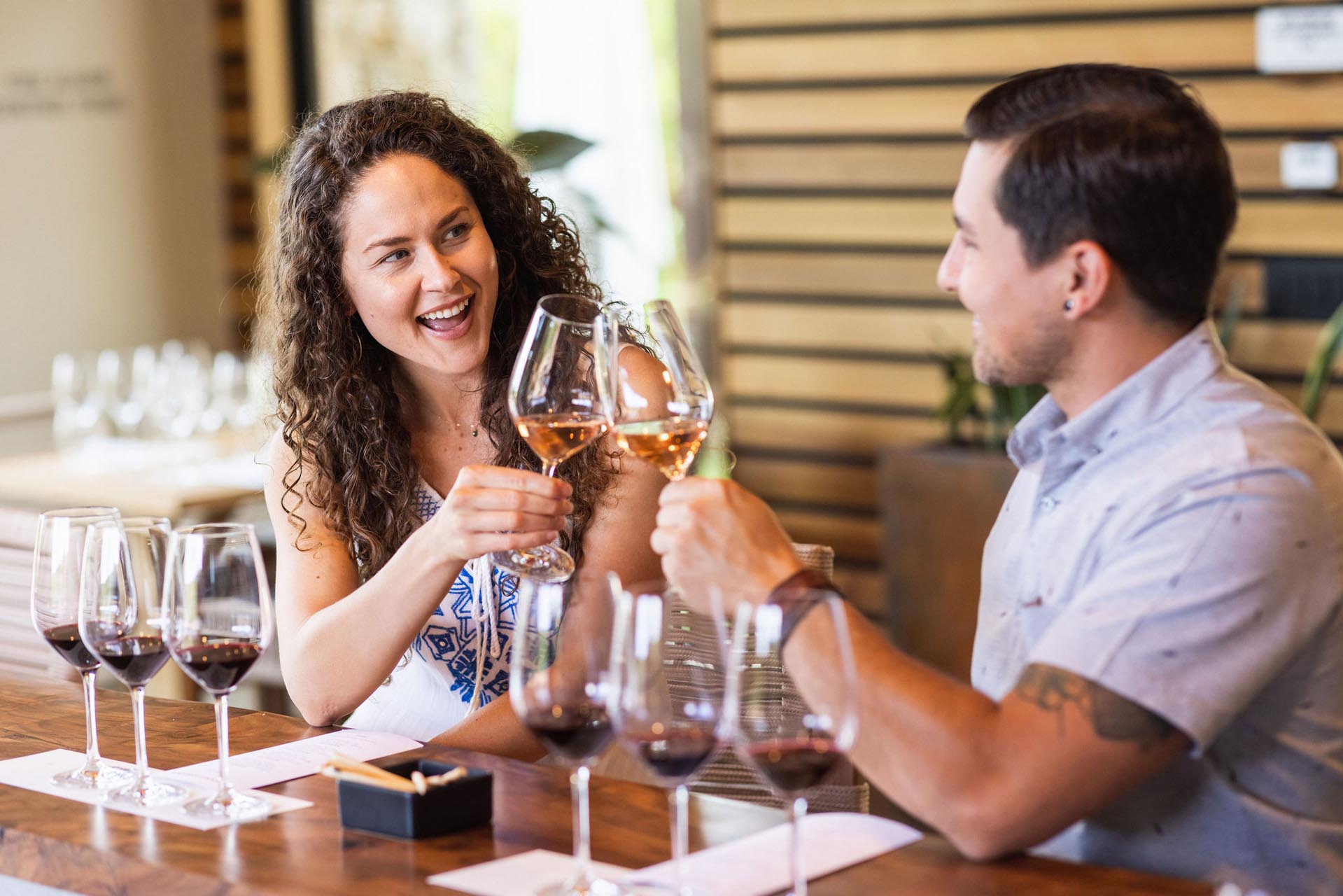 A woman and a man clink wine glasses while sitting at a wooden table with several glasses of red and white wine. They are smiling and appear to be enjoying their time together in a modern setting.