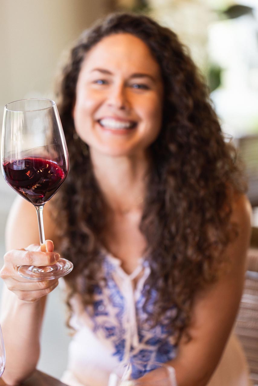 A smiling woman with long curly hair holds up a glass of red wine. She is wearing a sleeveless white top with blue patterns and is seated at a table. The background is softly blurred, suggesting a relaxed setting.