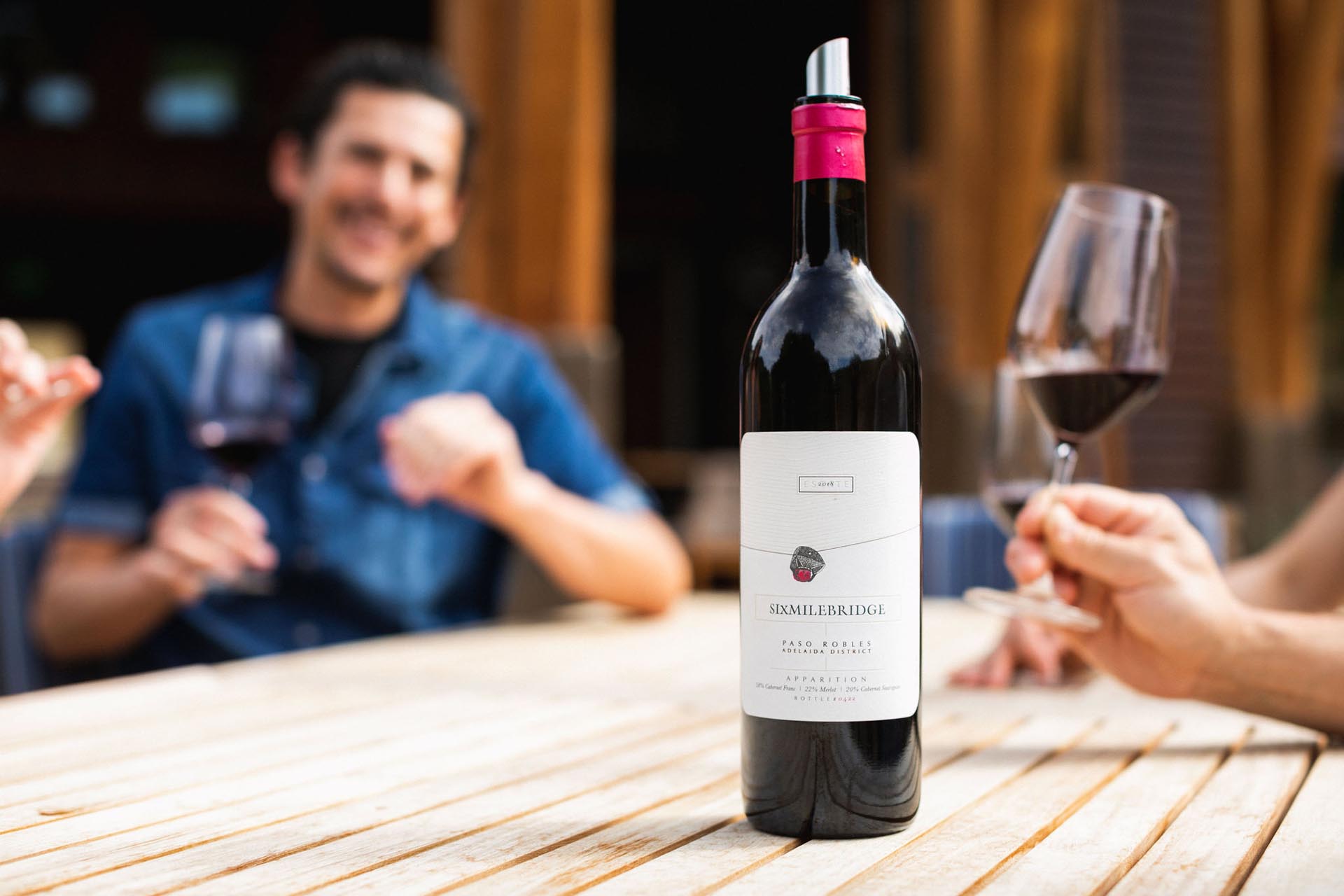 A bottle of wine with a red cap sits on a wooden table. Two people in the background hold wine glasses, engaged in conversation. The setting appears relaxed and outdoors.