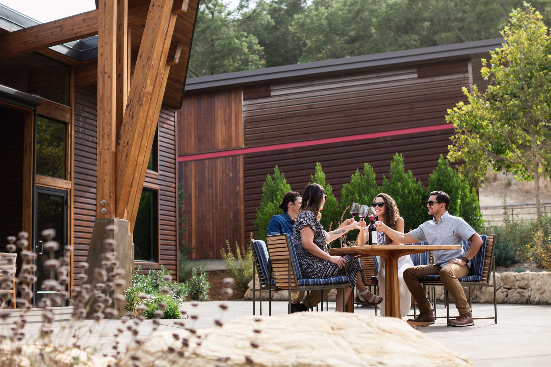 Four people sitting outdoors at a wooden table, toasting with glasses of red wine. They are on a patio surrounded by trees and modern wooden buildings. It is a sunny day, and they appear to be enjoying a social gathering.