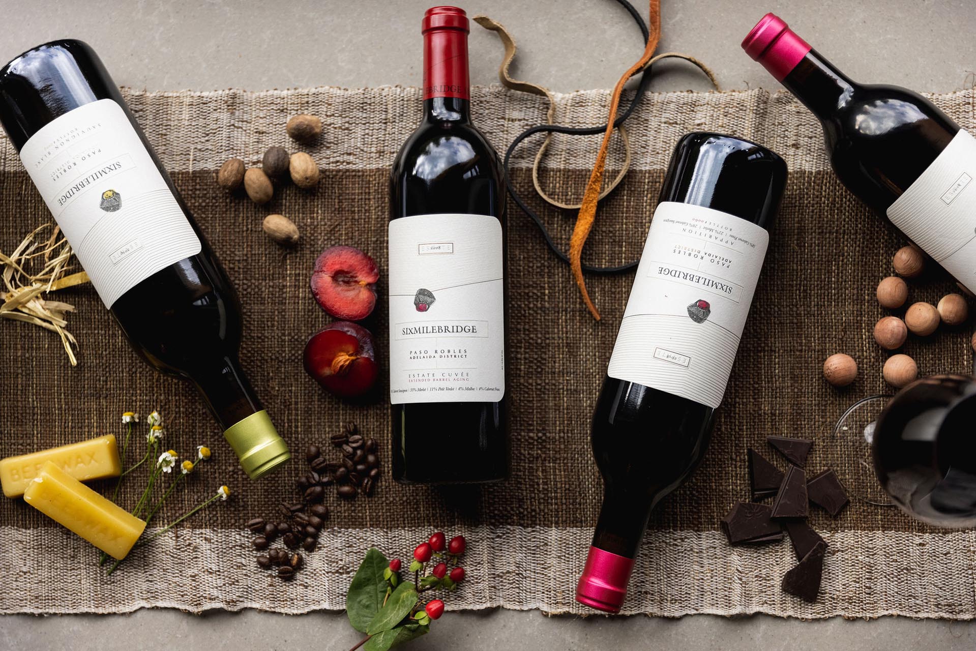 Four wine bottles are arranged on a textured brown cloth, surrounded by assorted items like a cut apple, nuts, chocolate, coffee beans, and herbs, creating a rustic and elegant display.