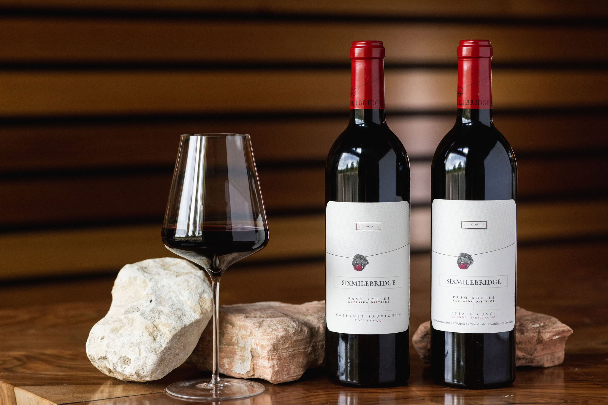 Two bottles of Sixmilebridge red wine with labels and red caps are placed beside a wine glass filled with red wine. The setup includes decorative stones, and the background features horizontal wooden panels.