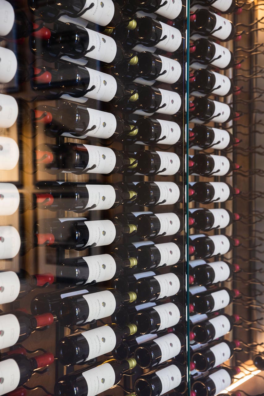 A display of wine bottles arranged on a wooden rack, each with a white label. Different cap colors indicate varying types or brands. Warm lighting creates a cozy ambiance.