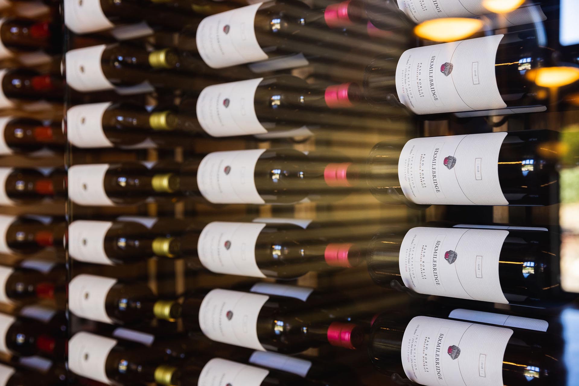 A display of wine bottles arranged on a wooden rack, each with a white label. Different cap colors indicate varying types or brands. Warm lighting creates a cozy ambiance.