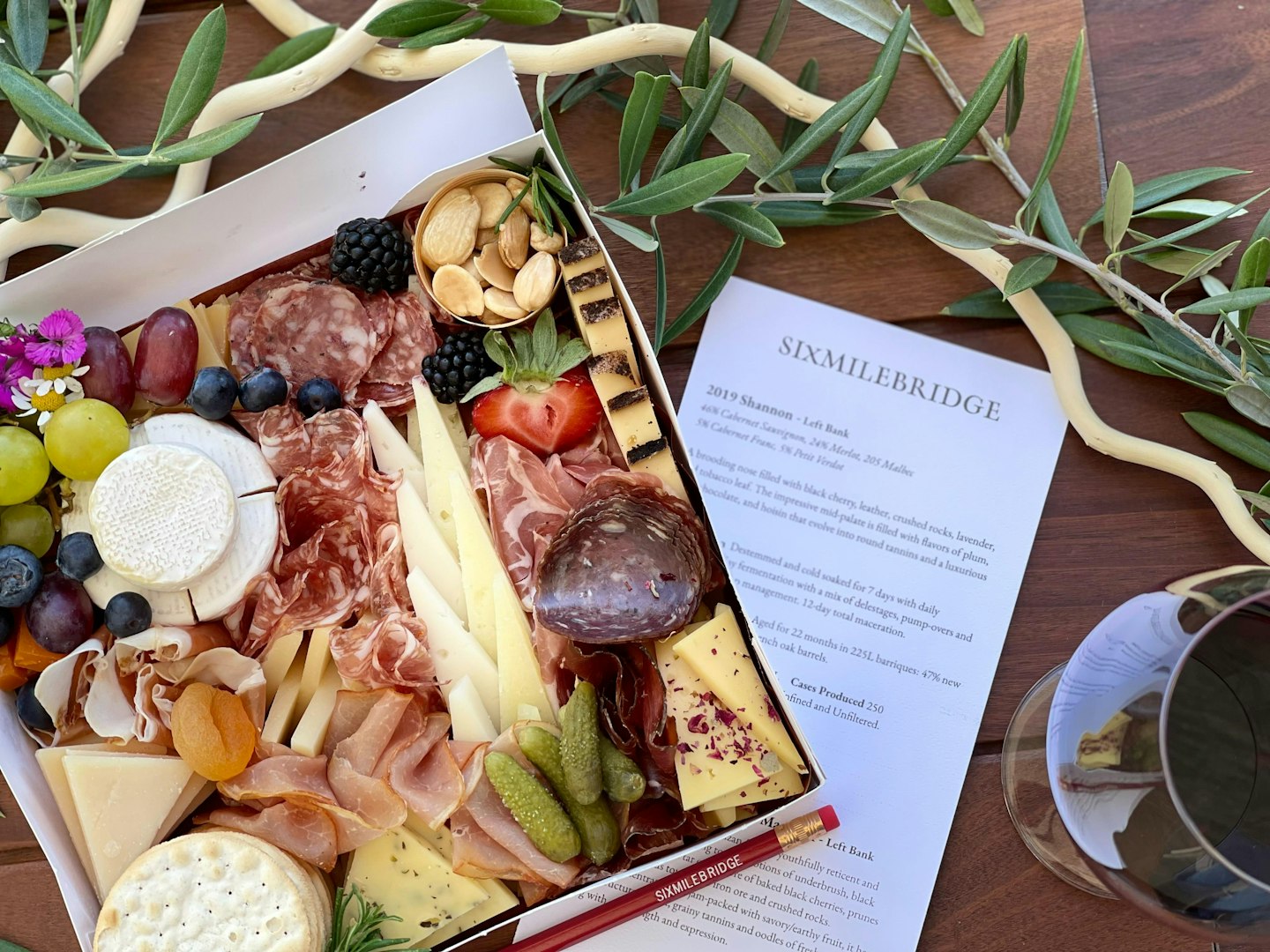 A box filled with cheese, meat, and fruit displayed on a table, showcasing a delightful assortment of fresh ingredients. This is underlaid by a Sixmilebridge wine tasting menu.